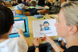 Foto: Kind mit Behinderung bedient Tablet und Frau sitzt neben ihm und hält das Symbol